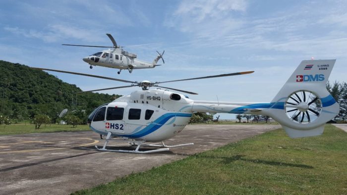 One helicopter on a concrete helipad surrounded by mountains and another helicopter above in mid flight.