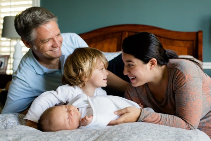 A husband and wife on their bed with their two children. They're all looking at each other, smiling.