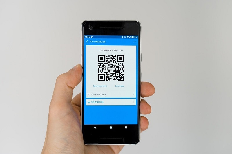 A hand holding a phone with the Alipay payment QR code on the screen. 