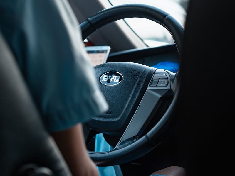 A photo of the steering wheel inside a  BYD brand Chinese car with the shoulder of a driver in the shot. 