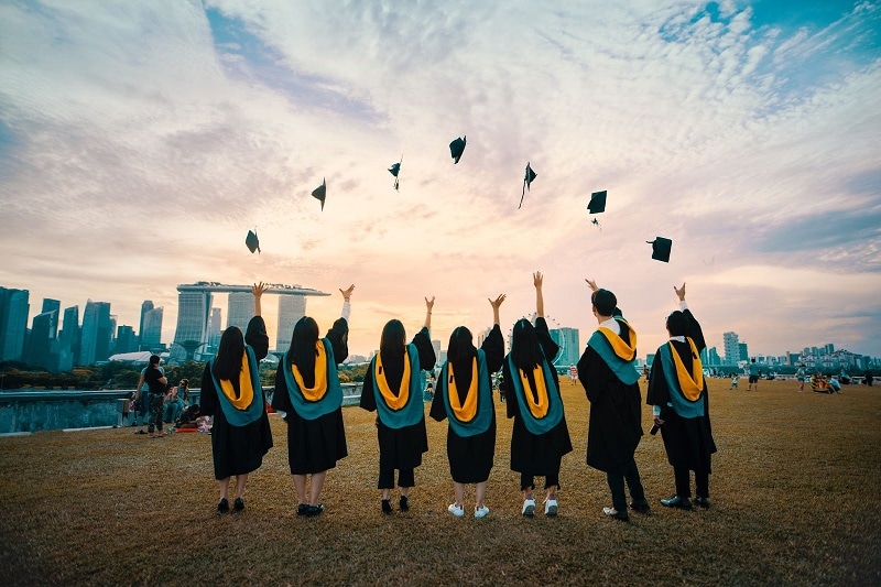 Graduating students throwing their caps in the air in a park with the Singapore skyline in the distance.