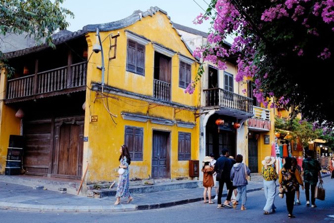 Hoi An street scene with old yellow houses, people wandering, and a purple flowering tree. 