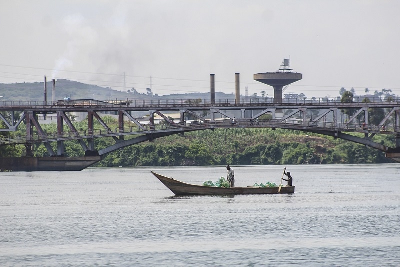 A small boat on a large river in Kampala Uganda. Bridge in the background and two men paddling the boat. 