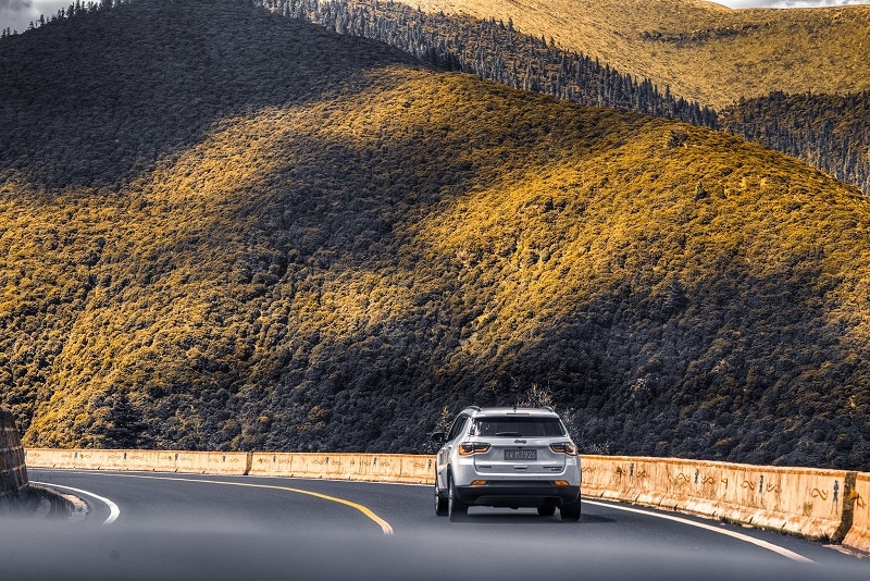 A photo of a white SUV with a Chinese license plate on a winding road with tree-covered hills all around. 